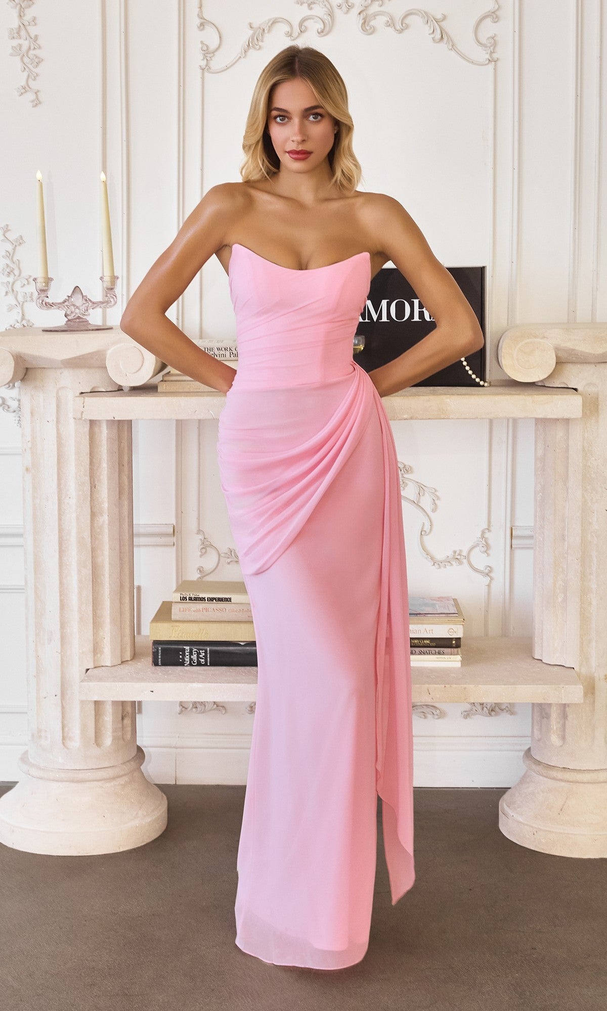 A woman stands indoors in front of a decorative white wall, two columns, and a shelf with books and candles, wearing the Amoris T726 Long Prom Dress in light pink with a strapless design, chiffon fabric, draping, and a lace-up back.